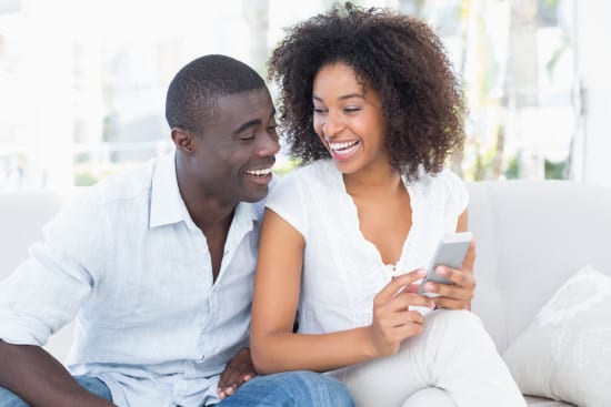 Attractive couple sitting on couch together looking at smartphone at home in the living room